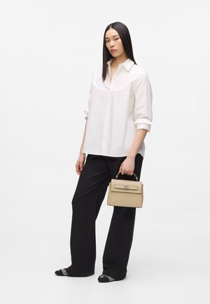 Ženska s ravnimi črnimi lasmi v beli srajci z gumbi, črnih širokih hlačah in črnih škornjih s koničastim nosom, v roki drži bež torbico za ročaj.
