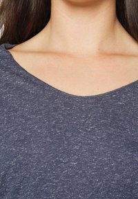Nahaufnahme des Halses und des Schlüsselbeins einer Person, die ein meliertes blaues Rundhalsshirt trägt, mit langen dunklen Haaren, die an den Seiten sichtbar sind.