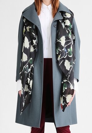 Manteau gris avec une grande écharpe fleurie noire. Le manteau présente un large col et une fermeture à boutons, tandis que l'écharpe arbore des motifs floraux blancs et verts.