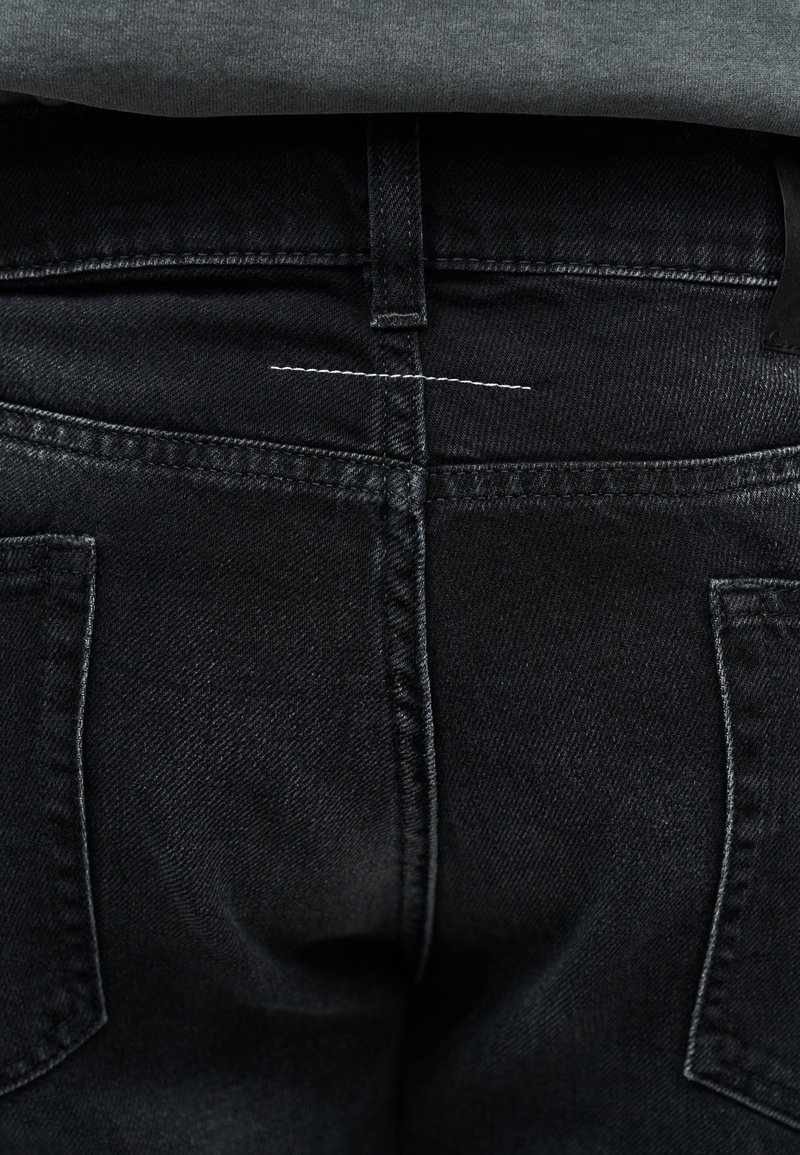 Primer plano de la parte trasera de unos vaqueros de mezclilla negros con presilla para cinturón visible, bolsillos traseros y una pequeña costura horizontal blanca encima de la costura.