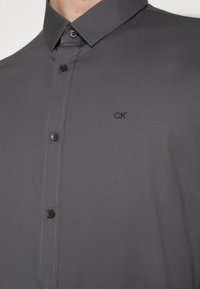 Donkergrijze blouse met kraag en geborduurd "CK" logo op de linkerborst.