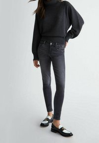 Femme portant un pull à col roulé sombre, un jean skinny noir délavé, et des mocassins noir et blanc avec un emblème doré, debout sur un fond clair.