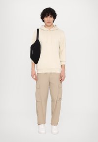 Felpa beige con tasca frontale, pantaloni cargo beige e sneakers bianche, abbinati a una borsa a tracolla nera con zip.