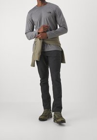 Graues Langarmshirt mit kleinem Logo, dunkle Hose und grüne Wanderschuhe. Inklusive einem olivfarbenen Sweatshirt, das um die Taille gebunden ist.