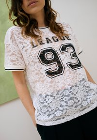 Femme portant une chemise en dentelle blanche à motifs floraux avec des bordures noires, inscription "LEAGUE 93" et pantalon noir, debout à l'intérieur les mains dans les poches.