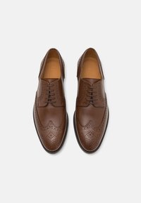 Scarpe in pelle marrone con lacci, design con perforazioni a punta ad ala, punta curva e superficie liscia. Presentano cuciture tono su tono e suola in gomma.