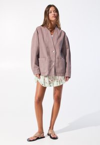 Licht mauve oversized jas met twee voorkruiszakken, sluiting met één knop en een ontspannen pasvorm, gedragen over een groene bloemenrok en sandalen.