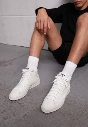 Person sidder på gulvet iført hvide høje sneakers, hvide crew-strømper, sorte shorts og en sort langærmet trøje.