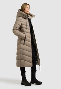 Donna che indossa un lungo cappotto imbottito beige con cappuccio, leggings neri e stivali neri alla caviglia, in piedi di lato con le mani in tasca.