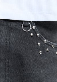 Gros plan sur un jean en denim noir avec des rivets argentés et une boucle anneau en D argentée, accompagné d'un haut en tissu côtelé blanc.