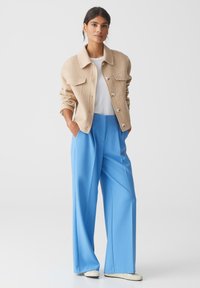 Beige wollen jas met knoopsluiting en twee borstzakken, gecombineerd met high-waisted, brede blauwe broeken. Witte shirt eronder.