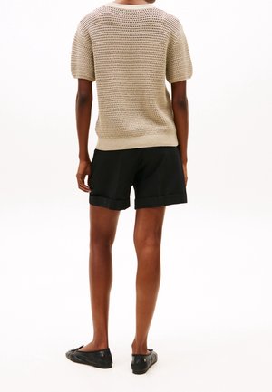 Personne debout de dos, portant un haut en tricot beige à manches courtes, un short noir retroussé et des ballerines noires sur fond blanc.