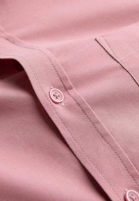 Roze katoenen shirt met knoopsluiting, opvallend gestikte naden en een gladde textuur. De knoop is lichtroze met vier gaten.