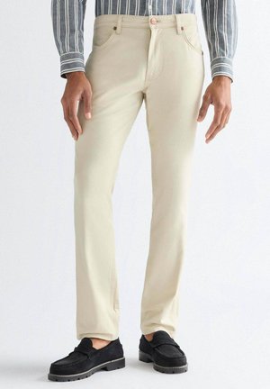 Mężczyzna w kremowych, prostych spodniach, czarnych mokasynach i niebieskiej koszuli w paski z długim rękawem stojący na jednolitym tle.