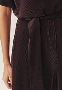 Örömteli pliszírozott ruha mély bordó színű anyagból, amelyet egy deréknál megkötött selyemöv díszít, és texturált függőleges minták találhatóak rajta.