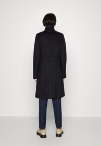 Abrigo negro ajustado con un cuello alto, cintura ceñida con cinturón y corte recto, combinado con pantalones azul marino y zapatos de color claro.