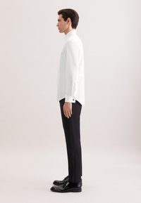 Mężczyzna stojący bokiem, ubrany w białą koszulę, czarne spodnie wizytowe oraz czarne, wypolerowane eleganckie buty na jednolitym jasnym tle.