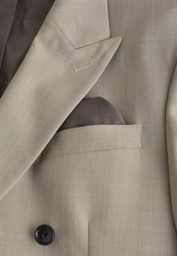 Blazer beige à col cranté, pochette sombre et détail de bouton noir sur tissu texturé.