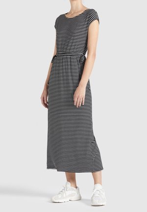Femme portant une robe midi à rayures horizontales noir et blanc avec des manches courtes et des baskets blanches, se tenant devant un fond uni.