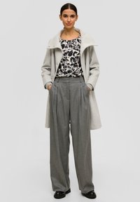 Hellgrauer Wollmantel mit hohem Kragen, kombiniert mit einem schwarz-weiß gemusterten Oberteil und grauen, plissierten Hosen. Schwarze Schuhe runden das Outfit ab.