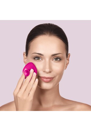 Vrouw met donker haar gebruikt een roze siliconen gezichtsreinigingsborstel op haar wang tegen een lichtpaarse achtergrond.