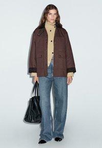 Giacca oversize marrone e testurizzata con colletto nero, dolcevita beige a costine, jeans blu a vita alta e borsa tote nera.