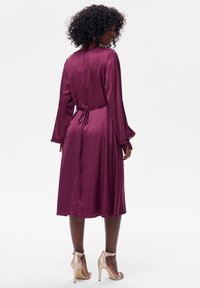 Satynowa sukienka w kolorze wina, z długimi, rozszerzanymi rękawami, paskiem w talii i długością do połowy łydki. Gładka faktura z subtelnym połyskiem oraz rozcięciem z tyłu.