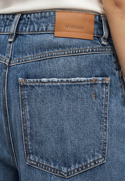 Κοντινό πλάνο πίσω τσέπης μπλε τζιν με εμφανή ραφές και καφέ δερμάτινη ετικέτα Sportmax στη ζώνη μέσης.