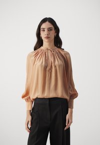 Blush silk blouse with a gathered neckline, drawstring closure, and billowy sleeves, paired with tailored black trousers.