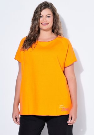 Donna sorridente con capelli mossi che indossa una maglietta larga arancione brillante con la scritta "Ho bisogno di Vitamina Mare", abbinata a pantaloni neri, in piedi contro un muro bianco.