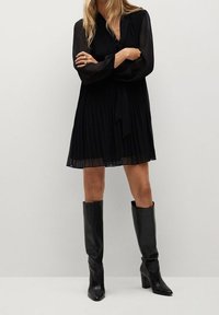 Vestido negro plisado con mangas largas translúcidas y un lazo en la cintura; combinado con botas altas de cuero negro con punta. 
