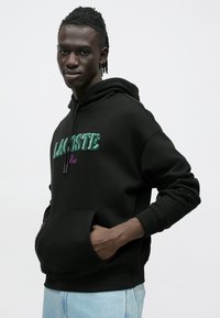 Czarna bluza z kapturem ze sznurkiem i przednią kieszenią. Na froncie wytłaczane zielono-fioletowe logo "LACOSTE Paris". Casualowy krój, miękki materiał.