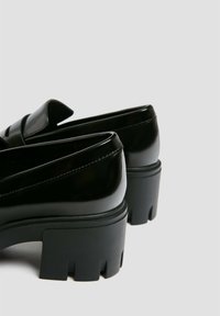 Mocassins à plateforme noire avec semelles épaisses rainurées, matériau brillant et talons bas, vus de dos sur un fond gris clair.