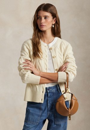 Ung kvinde med langt brunt hår har en cremefarvet kabelstrikket cardigan på, hvid skjorte, blå jeans og holder en tanfarvet halvmåneformet håndtaske.