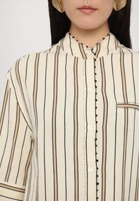Beige button-up shirt featuring vertical black and brown stripes, mandarin collar, and contrasting black stitching along the button placket.