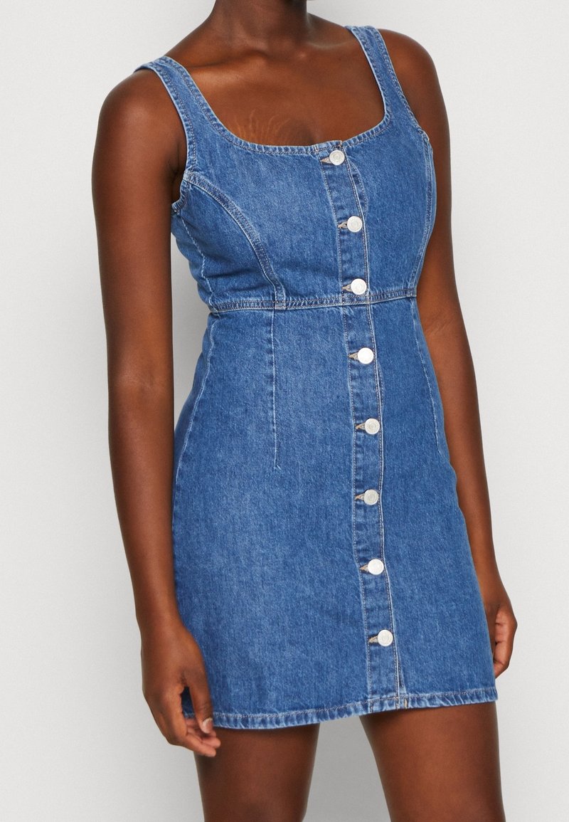 Nainen, jolla on päällään hihaton sininen denimpuku, jossa on edessä valkoiset napit, vyötäisillä tiukka ja joka päättyy polvien ylle.