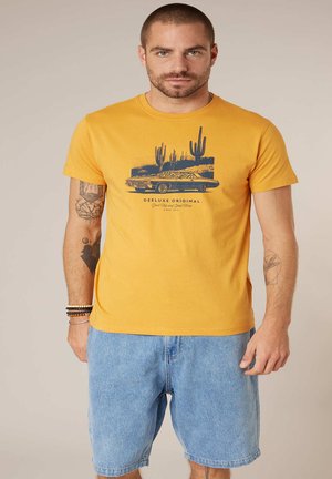 Man met een mosterdgele T-shirt met print van een cactus en een klassieker auto, gecombineerd met lichtblauwe denim shorts, staand tegen een beige achtergrond.