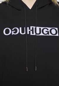 Černá mikina s kapucí a předním potiskem "ОГУHUGO" v kontrastní bílé. Obsahuje šňůrky a kovové konce. Měkká, strukturovaná tkanina.