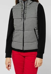 Schwarze und weiße Hahnentritt gepolsterte Weste mit hohem Kragen, schwarzen Ärmeln, zwei Seitentaschen und Kordelzug-Akzenten. Rote Hosen sichtbar.