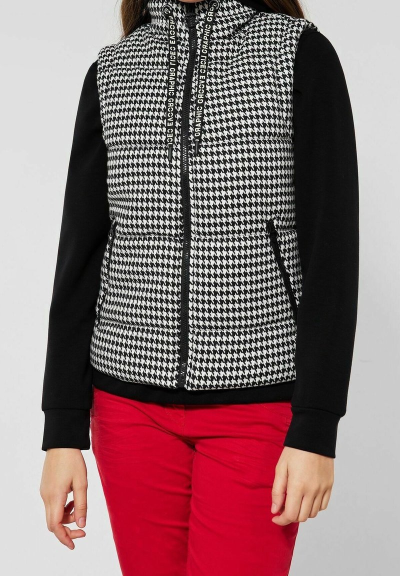 Schwarze und weiße Hahnentritt gepolsterte Weste mit hohem Kragen, schwarzen Ärmeln, zwei Seitentaschen und Kordelzug-Akzenten. Rote Hosen sichtbar.