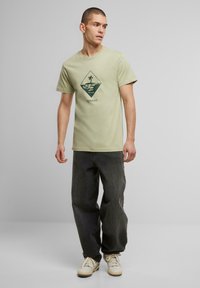 Camiseta de algodón de color verde claro con mangas cortas, que presenta un gráfico central de un diamante con una palmera y agua, combinada con unos jeans de color gris oscuro.
