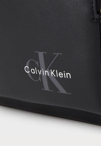 Črna torba iz sintetične usnje z izstopajočim srebrnim logotipom "Calvin Klein" v malih črkah in stiliziranim dizajnom "CK".