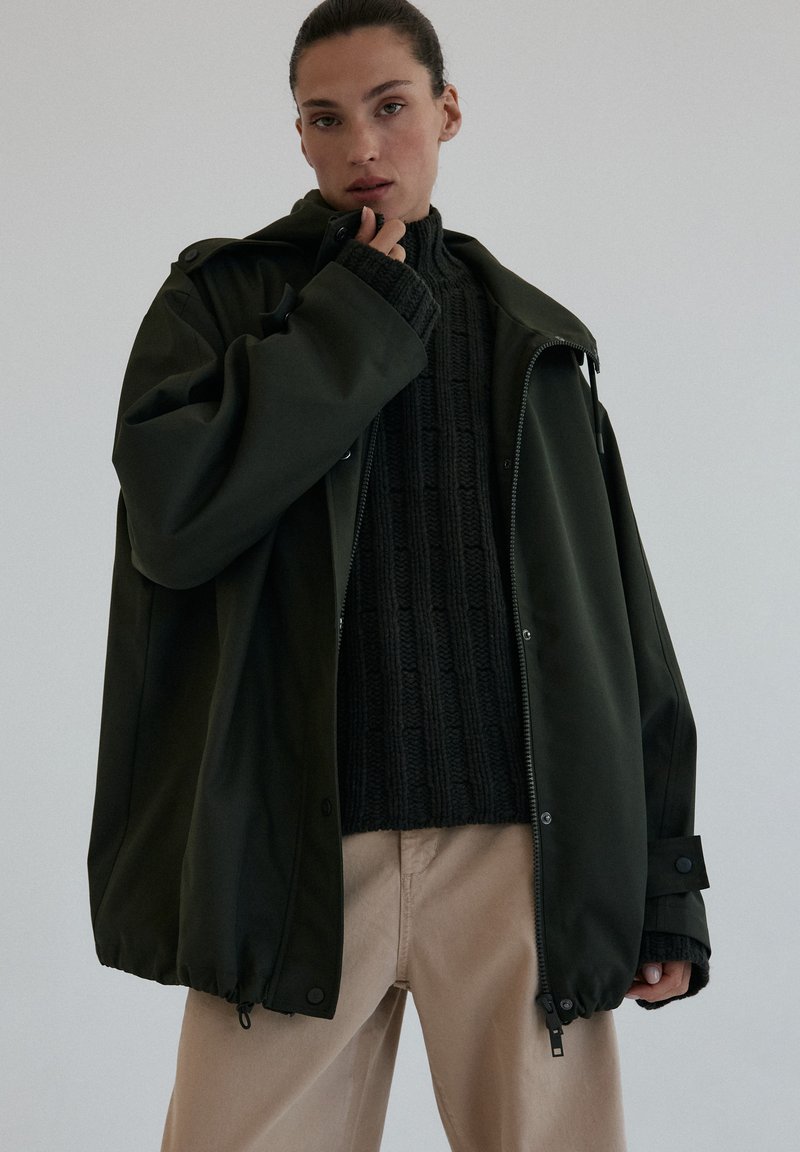 Przesadzona ciemnozielona wodoodporna kurtka z kapturem, zapinana na zamek, z elastycznymi mankietami, nałożona na teksturowany czarny sweter z dzianiny.