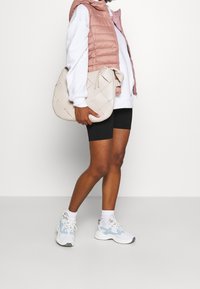 Gilet rembourré rose clair sur un sweat à capuche blanc, associé à un short noir et des baskets blanches avec des accents bleus. Sac en toile beige porté à la main.