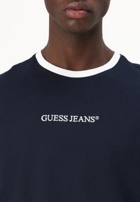 Camiseta azul marino con cuello de canalé blanco, que presenta el logo bordado "GUESS JEANS®" en el frontal. Fabricada en suave material de algodón.