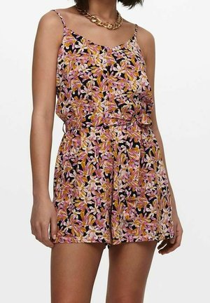 Femme portant une combinaison sans manches à fleurs roses, jaunes et blanches sur fond noir et un collier chaîne épais en or.