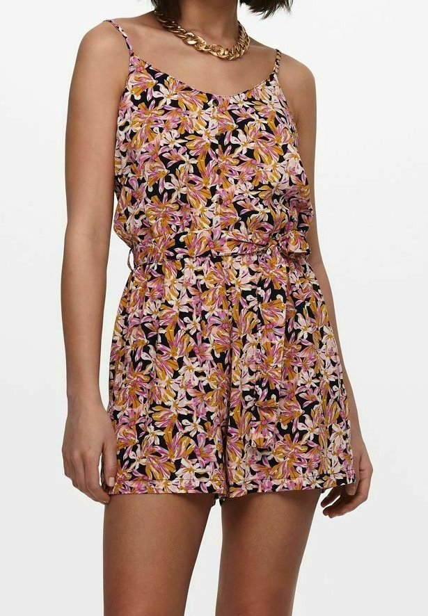 Femme portant une combinaison sans manches à fleurs roses, jaunes et blanches sur fond noir et un collier chaîne épais en or.