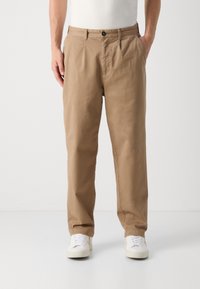 Hellbraune Baumwollhosen mit lockerer Passform, gefaltetem Vorderteil und seitlichen Taschen. Mit einem schwarzen Knopf an der Taille befestigt.