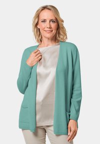 Türkiser Cardigan aus weichem Strickmaterial, mit offener Vorderseite, langen Ärmeln und zwei seitlichen Taschen. Tragen über einem leichten Oberteil.