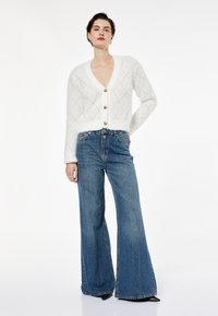 Biały puszysty kardigan w diamentowy wzór, wyposażony w duże złote guziki, zestawiony z dżinsami o fasonie flare w niebieskim kolorze. Gładka tekstura, luźny krój.